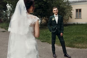 Handsome happy groom in stylish dark green wedding suit smiling and looking at beautiful bride in white wedding dress with bouquet, newlyweds posing in the park