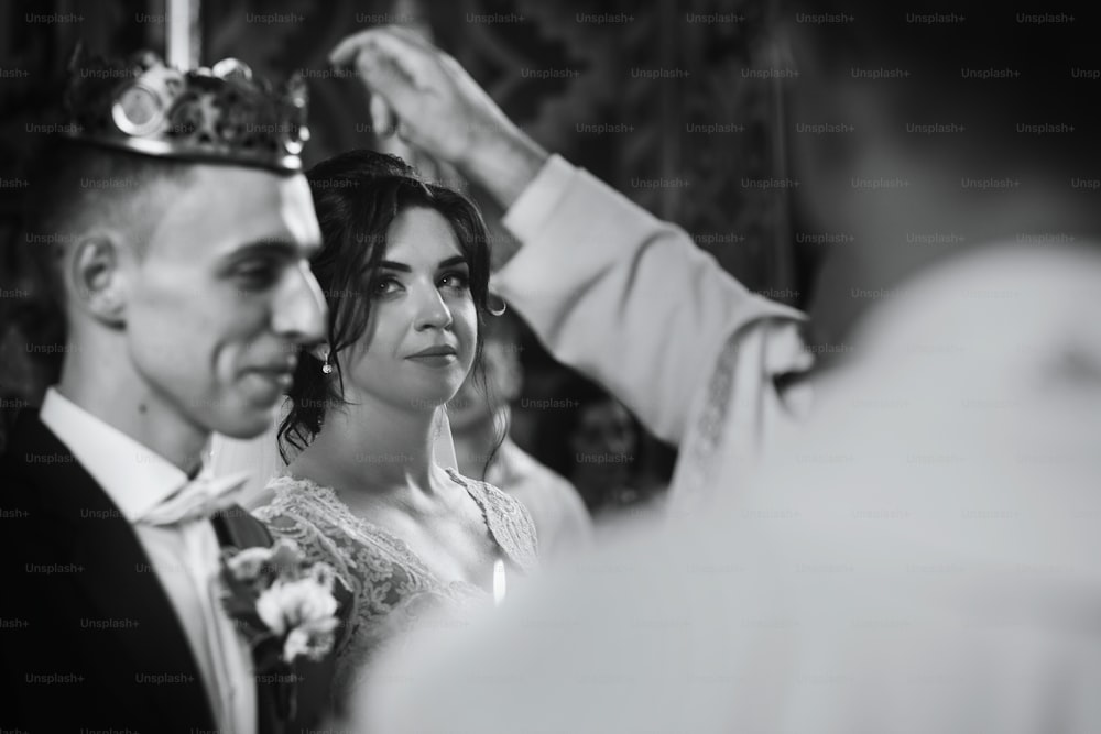 Feliz pareja espiritual, novio elegante y hermosa novia morena en vestido blanco sosteniendo velas en la ceremonia de la iglesia de la boda durante la coronación de la corona dorada