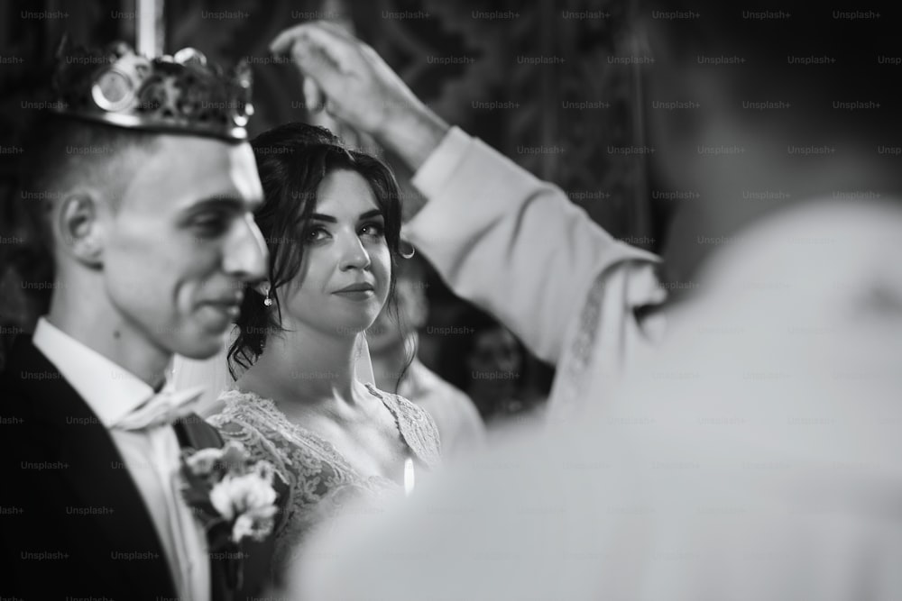 Happy spiritual couple, stylish groom and beautiful brunette bride in white dress holding candles at wedding church ceremony during golden crown coronation