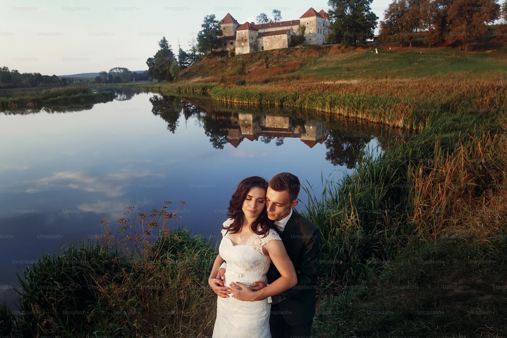 Romantic newlywed couple hugging near blue lake, sensual groom embracing gorgeous bride from behind near river, castle in the background, sky and castle reflection in the water