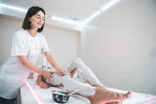 Enjoying procedure. Cheerful beautician in white lab coat gently touching woman hip and knee covered with cosmetic mask. Young lady in white underwear lying on massage table