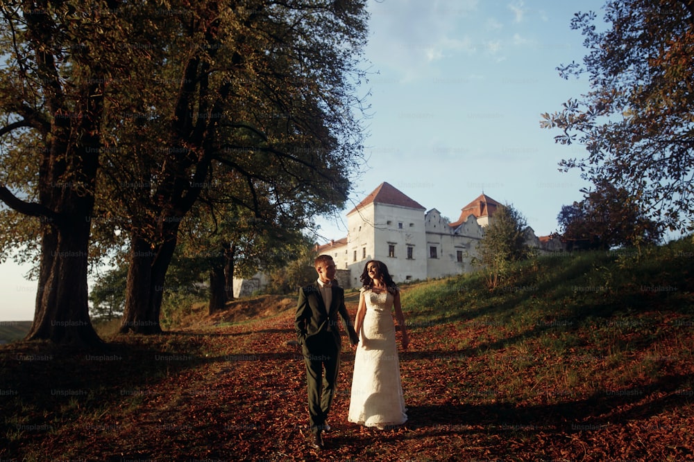 luxury wedding couple  walking holding hands in soft evening light at sunset near castle. sensual romantic moment of beautiful gorgeous bride and stylish groom outdoors