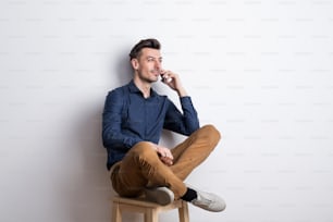 スタジオのスツールに座って電話をかけている紺色のシャツを着てスマートフォンを持っている若いハンサムな男性の肖像画。