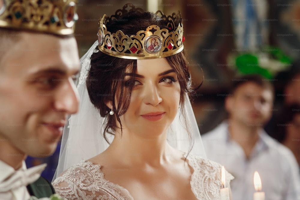 Gorgeous brunette bride in golden crown and vintage white dress looking at happy emotional groom during wedding ceremony in church, face closeup