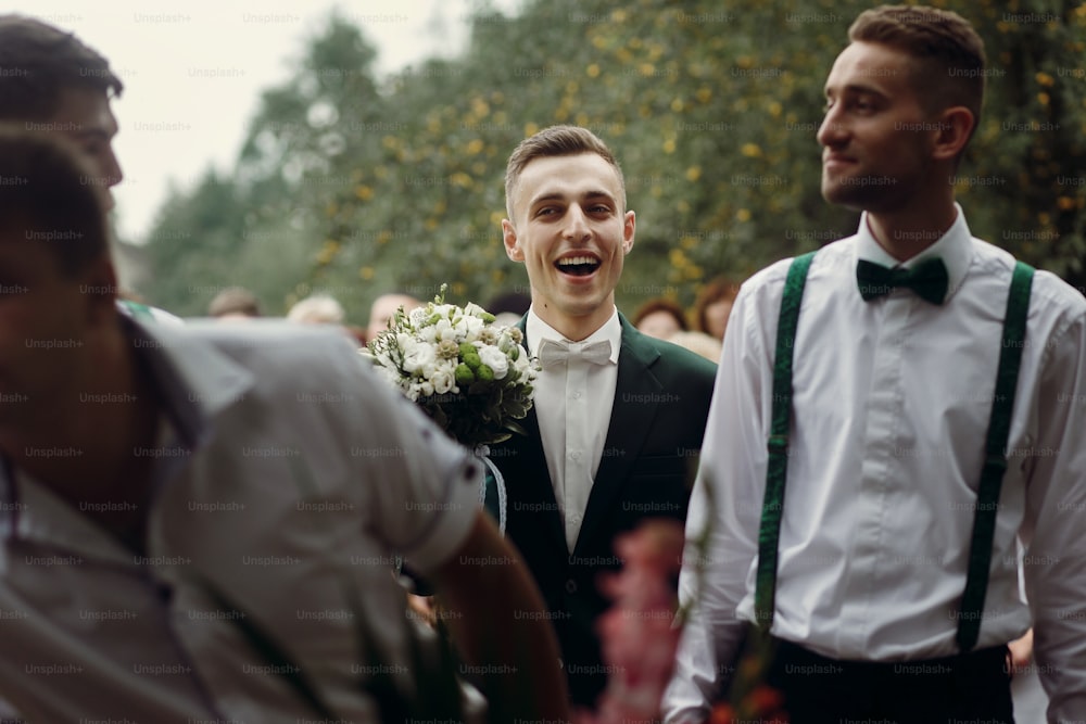 Handsome happy brunette groom in stylish black suit with white roses wedding bouquet walking to office for ceremony with guests and groomsmen