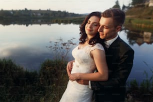 Romantic newlywed couple hugging near blue lake, sensual groom embracing gorgeous bride from behind near river, castle in the background, sky and castle reflection in the water