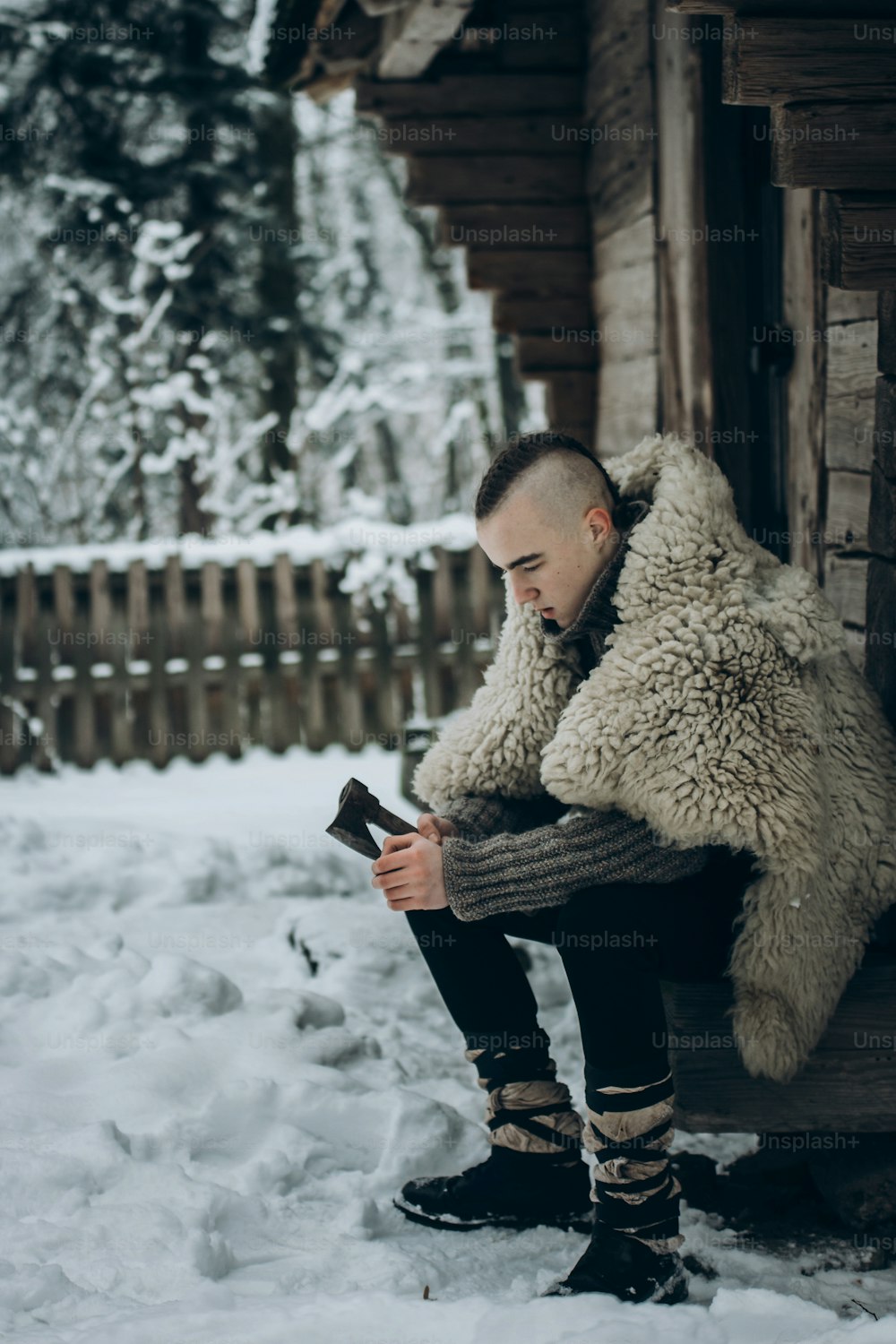 strong viking warrior sharpening his weapon near wooden hut in scandinavian forest in the winter, thor cosplay costume, northern lumberjack with mohawk hairstyle sitting with axe, viking concept