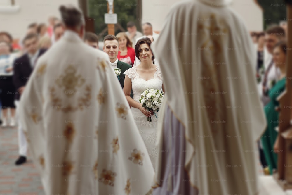 Glückliches, emotionales Paar, Braut in weißem Kleid und Blumenstrauß und hübscher Bräutigam, der zur christlichen Hochzeitszeremonie im Freien in Richtung Priester geht
