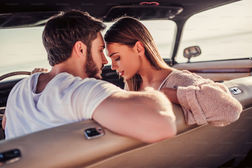 La pareja romántica está sentada en un auto retro verde en la playa. Hombre guapo barbudo y atractiva joven en coche clásico antiguo. Elegante historia de amor. Abrazarse y besarse mientras se está en el coche.