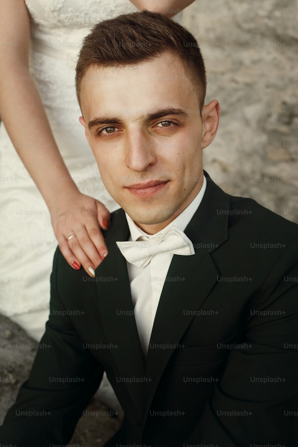 Handsome confident groom in stylish dark green suit with white bowtie sitting on stone steps near beautiful bride in white dress near castle entrance, romantic portrait