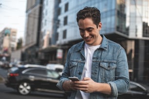 Waist up portrait of smiling young male standing on street with phone in hands. He is surfing net while waiting for date with delight