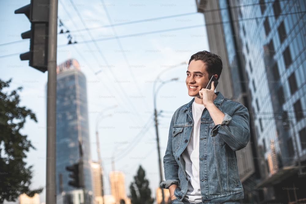 I am waiting. Waist up portrait of smiling male walking in city and talking on mobile. He is looking sideways with delight while expecting date with girlfriend. Copy space in left side