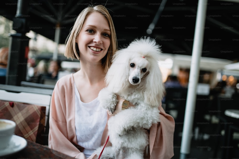 Mulher jovem bonita e elegante sentada no bar do café com seu lindo poodle anão branco e olhando para a câmera