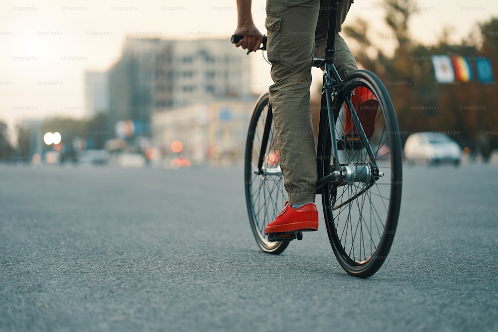 Close-up de pernas de homem casual andando de bicicleta clássica na estrada cinza da cidade vestindo tênis vermelhos e calças confortáveis. Espaço de cópia