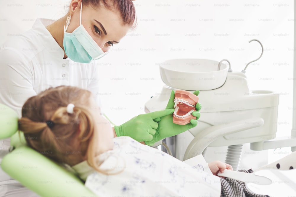 Little girl in dentists chair, being educated about proper tooth-brushing by her paediatric dentist. Early prevention and oral hygiene concept.