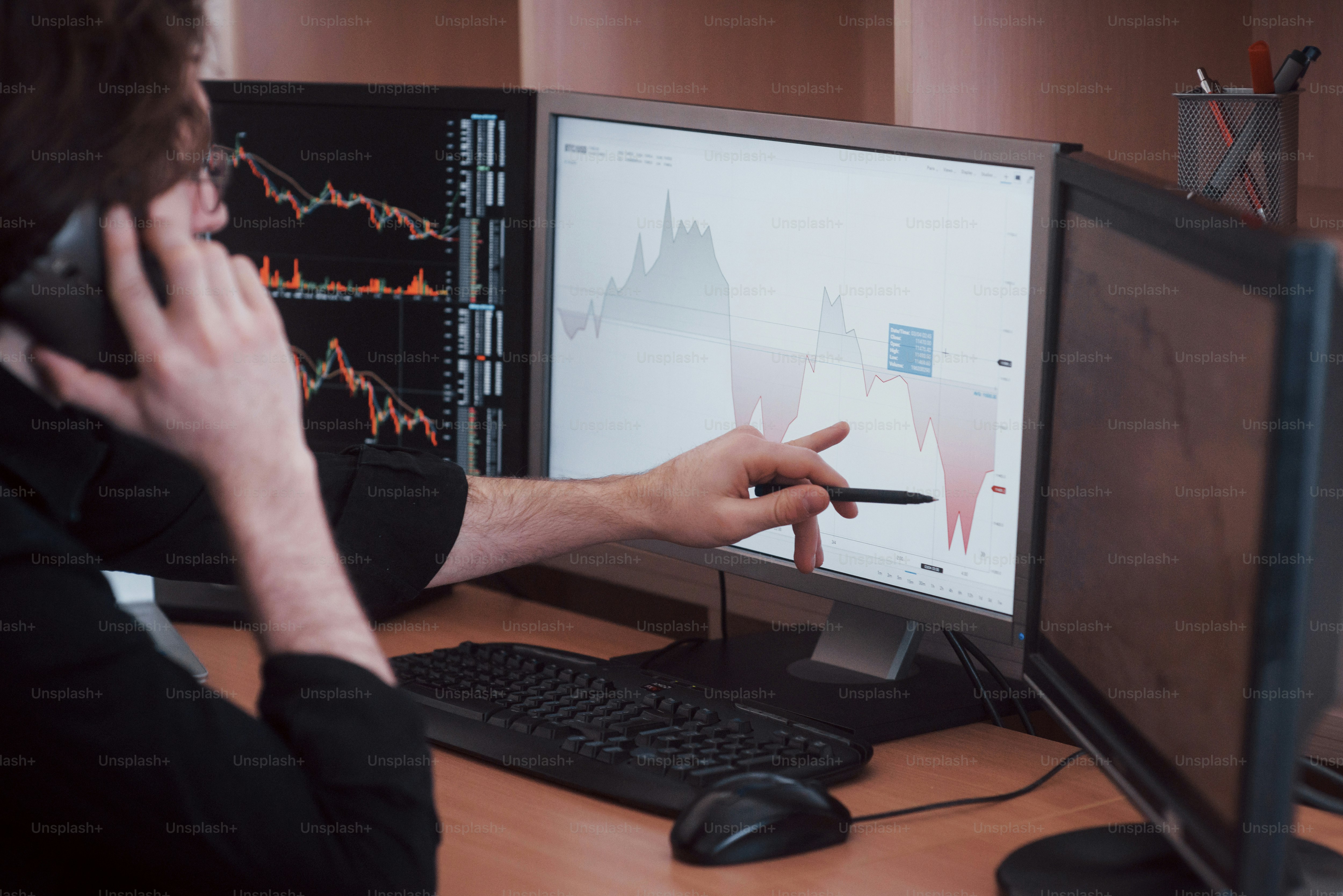 Over the shoulder view of and stock broker trading online while accepting orders by phone. Multiple computer screens ful of charts and data analyses in background.