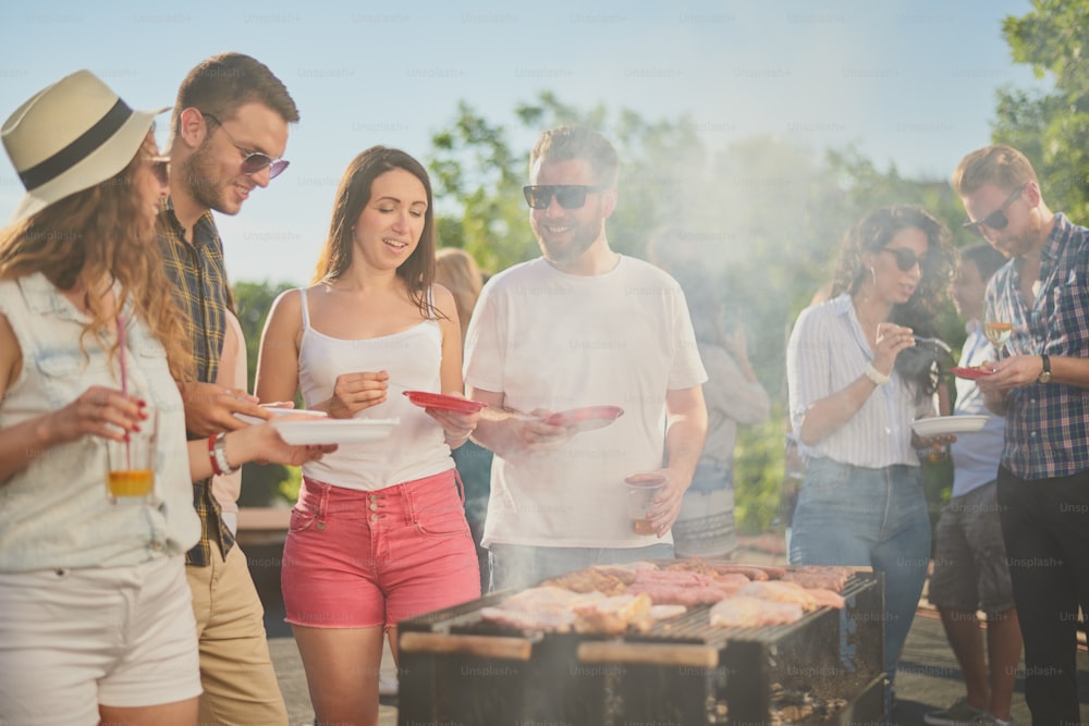 Group of people standing around grill, chatting, drinking and eating.