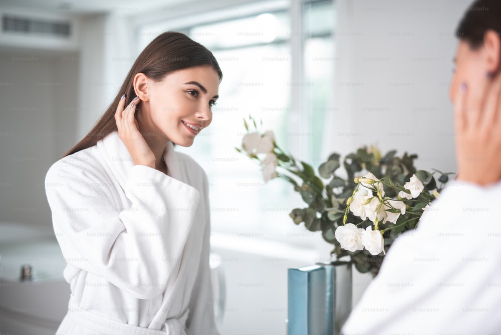 Portrait of outgoing young girl touching hair with arm while looking at mirror indoor. She wearing white bathrobe