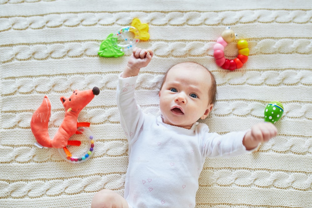 Newborn baby girl lying on knitted blanket, smiling and looking at colorful wooden rattle toys. 1 month old child at home