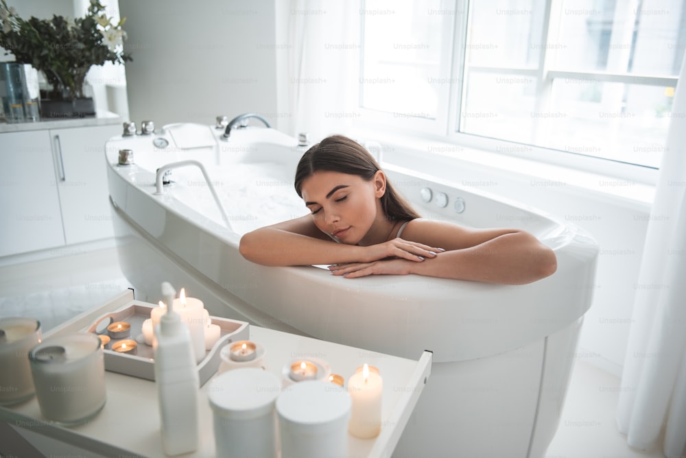 Portrait of calm girl with closing eyes having leisure in water with foam opposite candles in apartment