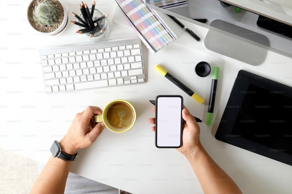 Diseñador gráfico sosteniendo un teléfono móvil con pantalla en blanco en el espacio de trabajo del artista creativo. Vista superior del lugar de trabajo
