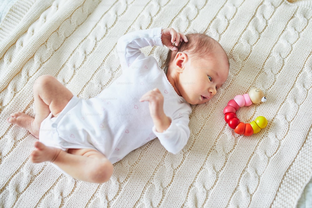 Newborn baby girl lying on knitted blanket, smiling and looking at colorful wooden toy. Three weeks child at home
