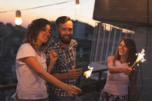 Group of young friends having fun at a rooftop party, singing, dancing and waving with sparklers. Focus on the guy