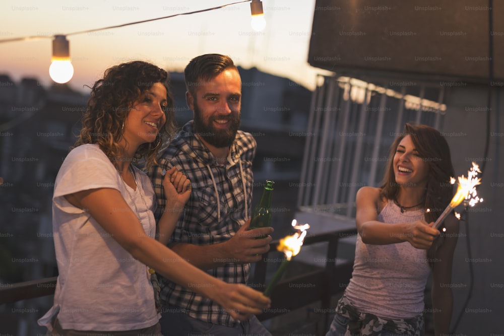 Group of young friends having fun at a rooftop party, singing, dancing and waving with sparklers. Focus on the guy