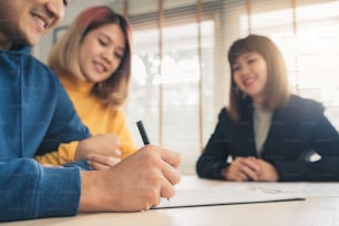 Happy young Asian couple and Real Estate Agent agent. Cheerful young man signing some documents while sitting at desk together with his wife. Buying new house real estate. Signing good condition contract.