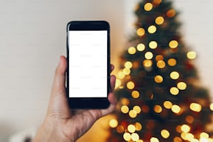 아름다운 크리스마스 트리 조명을 배경으로 빈 화면이 있는 휴대폰을 들고 있습니다. 메리 크리스마스와 새해 복 많이 받으세요. 텍스트 공간. 계절 인사, 행복한 휴일