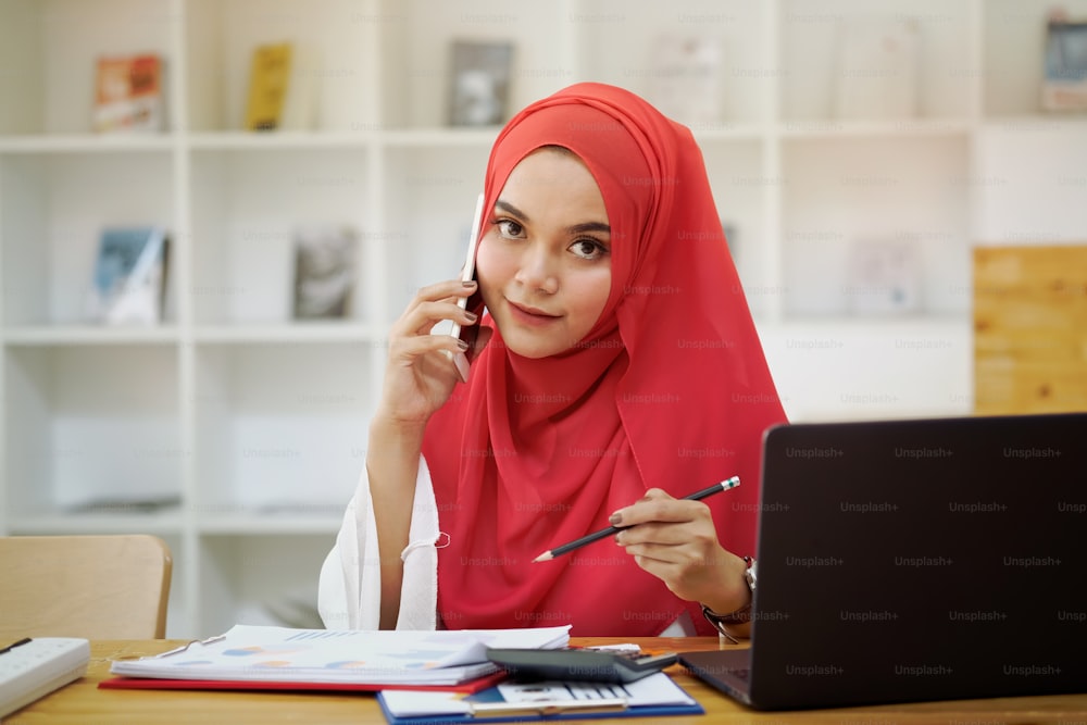 Junge muslimische Geschäftsfrau ruft ihr mobiles Smartphone am Schreibtisch an.