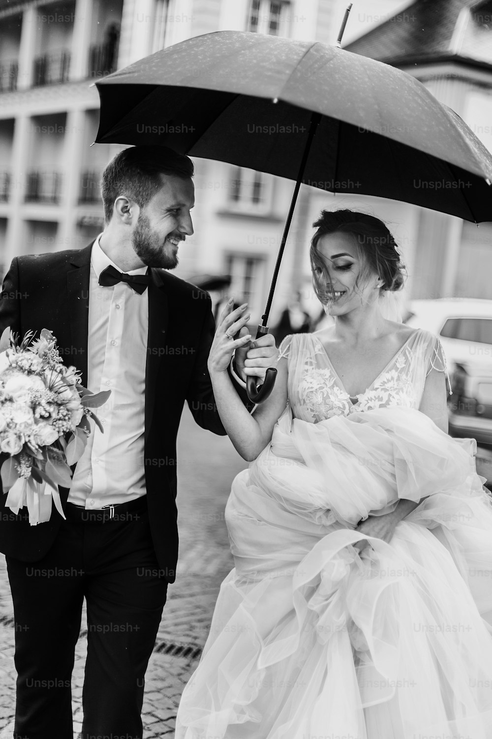 Wunderschöne Braut und stilvoller Bräutigam, die unter dem Regenschirm in einer regnerischen Straße spazieren gehen und lächeln. Sinnliches Hochzeitspaar, das sich umarmt. Romantische Momente des Brautpaares. Modernes Hochzeitsfoto