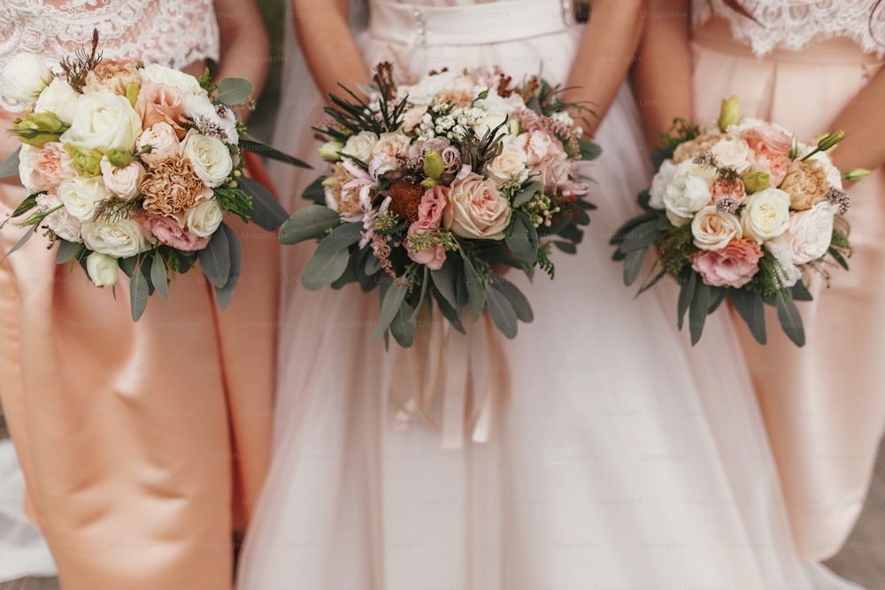 Splendida sposa e belle damigelle d'onore che tengono eleganti bouquet da sposa sullo sfondo di abiti moderni. Decorazioni per matrimoni