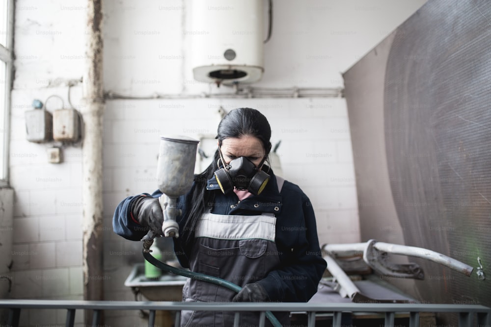 Mulher forte e digna fazendo um trabalho árduo. Ela usando compressor de pulverização industrial para pintar alguns produtos de metal.