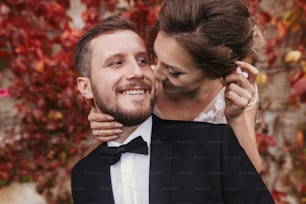 Gorgeous bride and stylish groom gently hugging and smiling at wall of autumn red leaves. Happy sensual wedding couple embracing. Romantic moments of newlywed