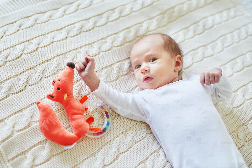 生まれたばかりの女の子の赤ちゃんがニットの毛布の上に横たわり、笑顔でカラフルな木製のガラガラのおもちゃを見ています。生後1ヶ月のお子さんがいらっしゃいます