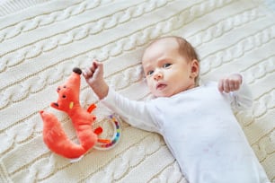 Newborn baby girl lying on knitted blanket, smiling and looking at colorful wooden rattle toy. 1 month old child at home