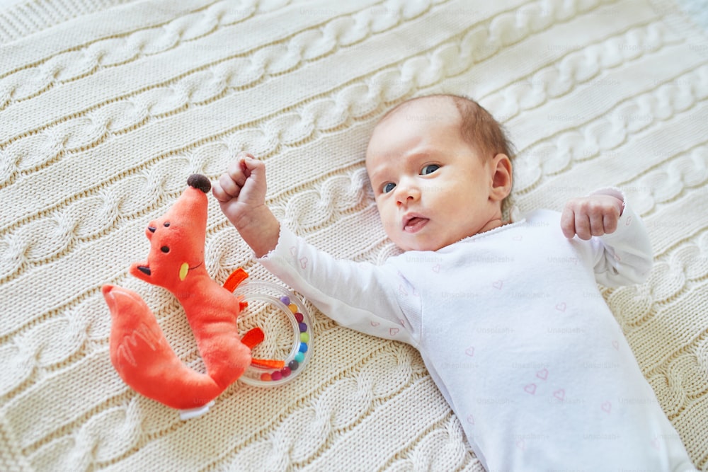 Newborn baby girl lying on knitted blanket, smiling and looking at colorful wooden rattle toy. 1 month old child at home