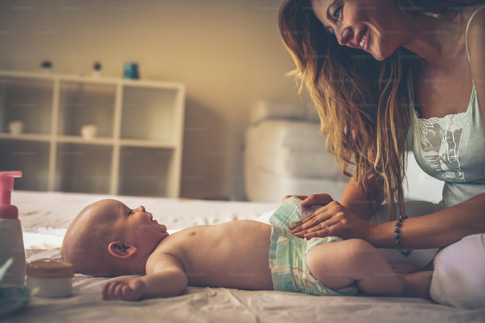 ベッドで女の赤ちゃんと遊ぶ若い母親。赤ちゃんのおむつを替えるお母さん。家族。