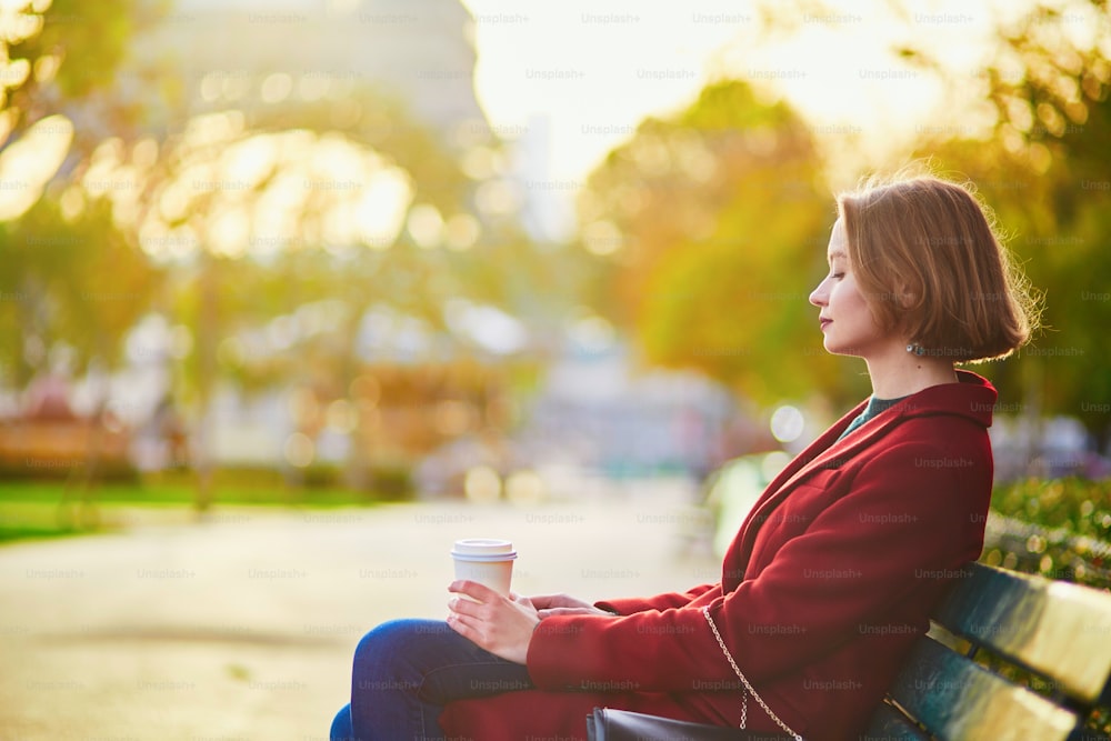 Beautiful young French woman sitting on the bench and drinking coffee near the Eiffel tower in Paris on a fall day