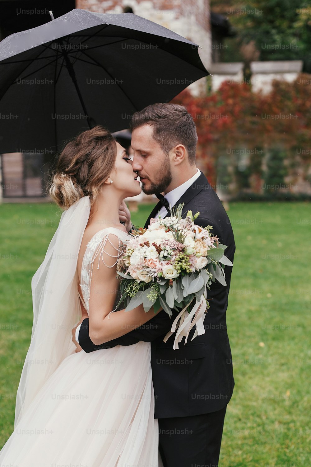 Wunderschöne Braut und stilvoller Bräutigam, die sich leidenschaftlich unter dem Regenschirm in regnerischer Natur küssen. Sinnliches Hochzeitspaar, das sich umarmt. Romantische Momente des Brautpaares. Modernes Hochzeitsfoto