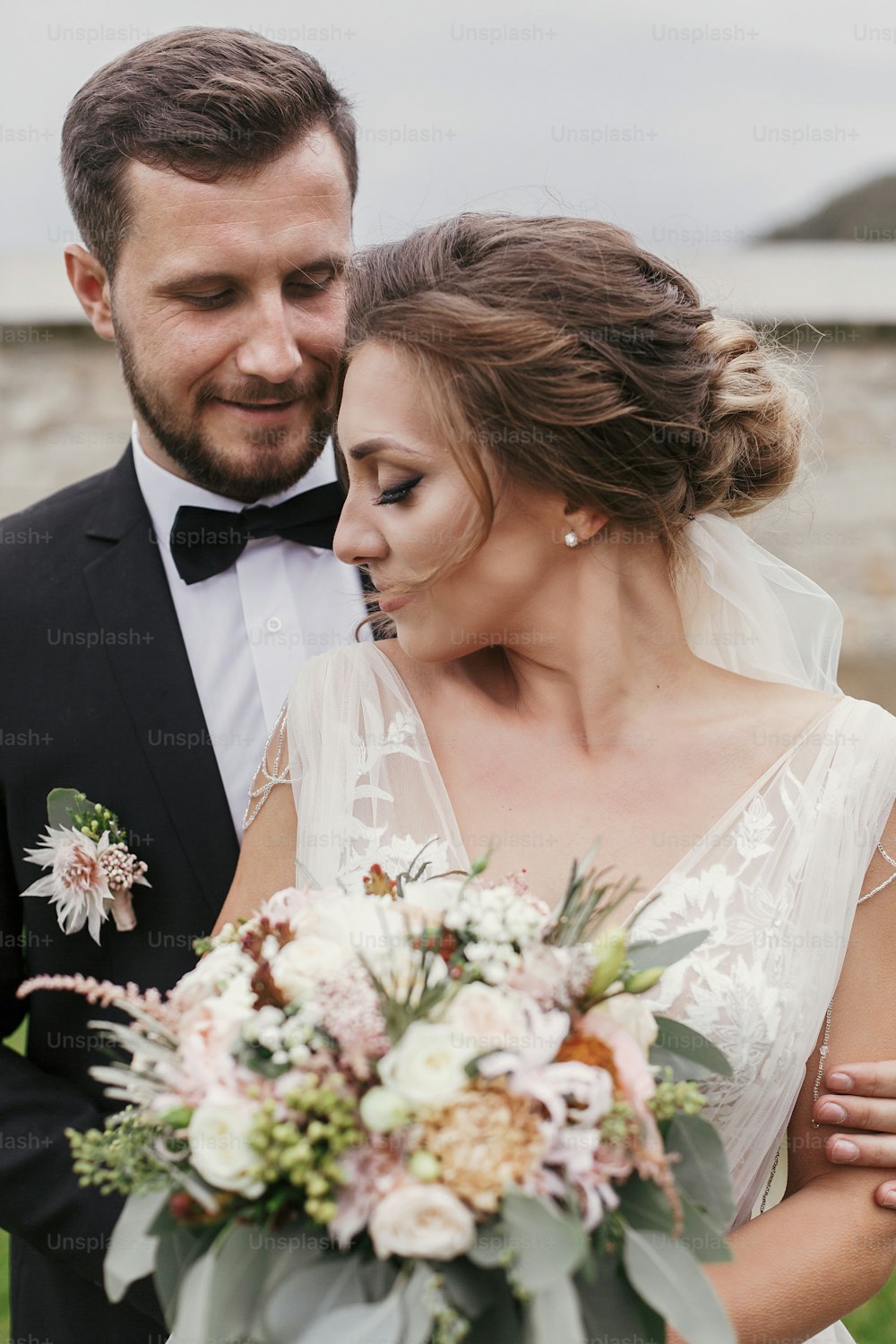 Gorgeous bride with modern bouquet and stylish groom gently hugging and smiling outdoors. Sensual wedding couple embracing. Romantic moments of newlyweds. Wedding photo
