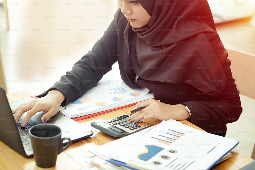 Jovem asiática atraente usando a papelada de trabalho escura do hijab com seu laptop.