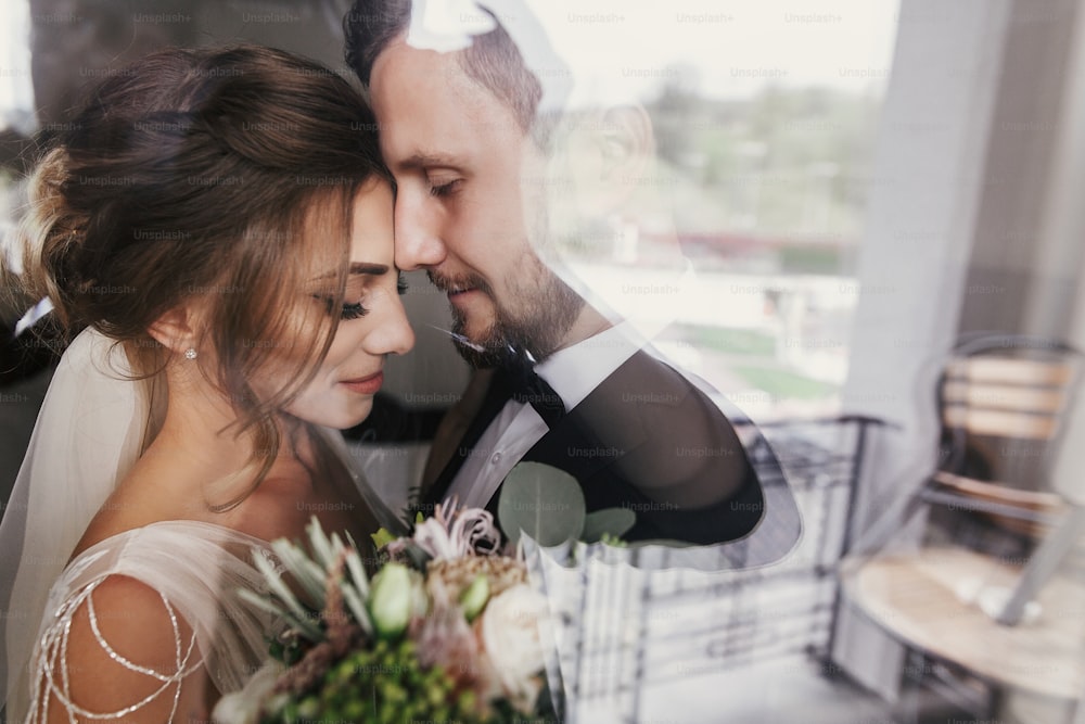 Gorgeous bride and stylish groom gently hugging at window. Sensual wedding couple embracing. Romantic moments of newlyweds. Creative wedding photo through glass. Copy space