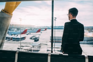 Back of a young business man standing with the suitcase at the airport waiting for the flight