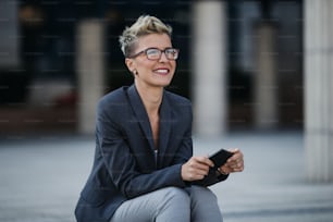Jeune femme d’affaires heureuse debout devant un grand bâtiment moderne. Elle sourit et parle sur son téléphone portable.
