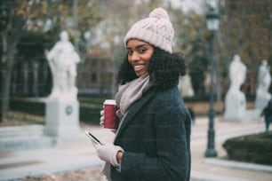 Joven mujer negra bebiendo café y hablando por teléfono móvil alegremente en invierno