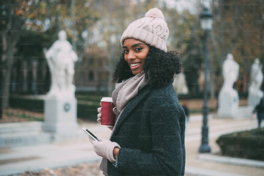 젊은 흑인 여성이 커피를 마시고 겨울에 유쾌하게 휴대 전화로 말한다