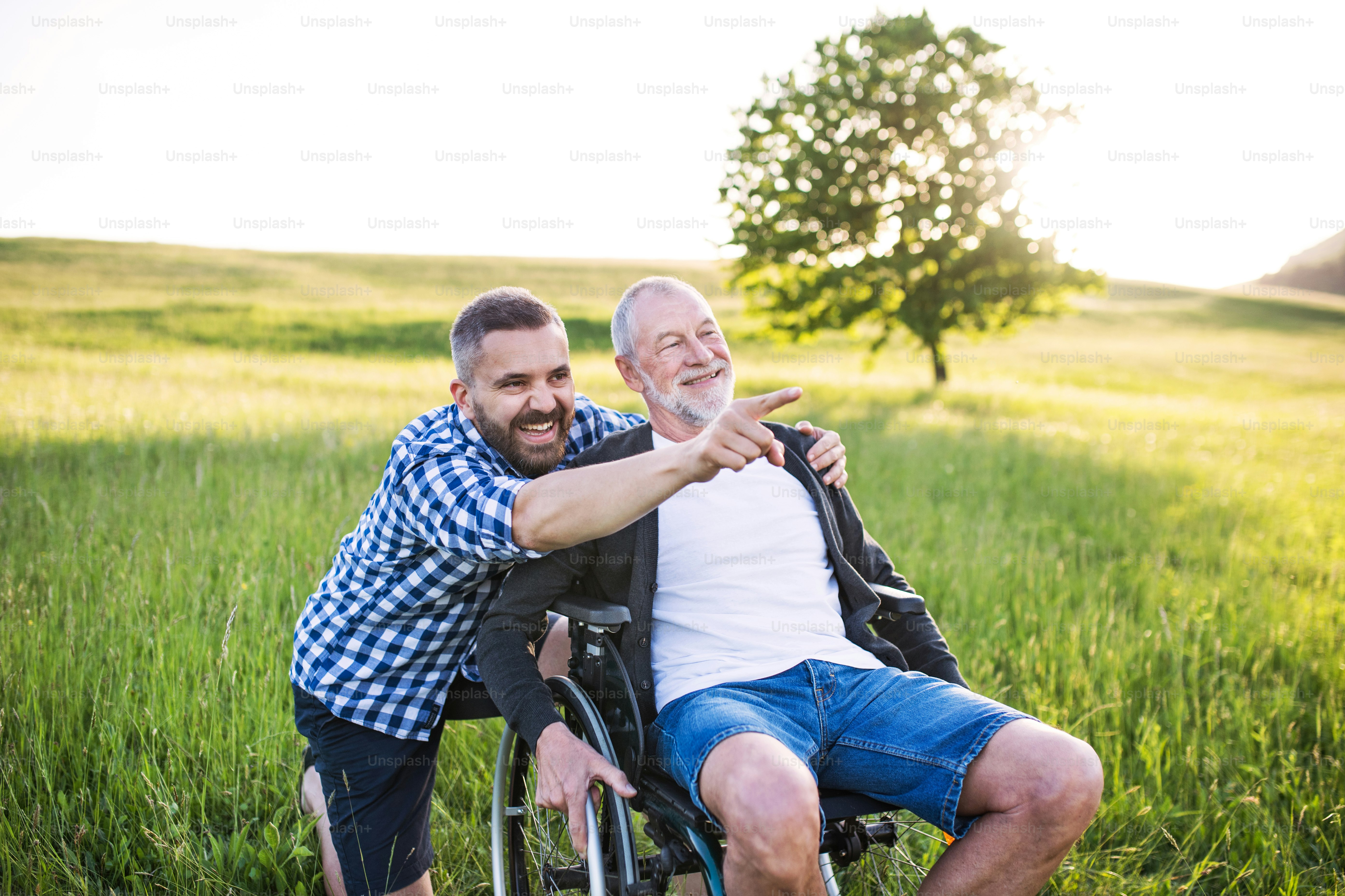 Un figlio hipster adulto con il padre anziano in sedia a rotelle durante una passeggiata su un prato nella natura al tramonto.