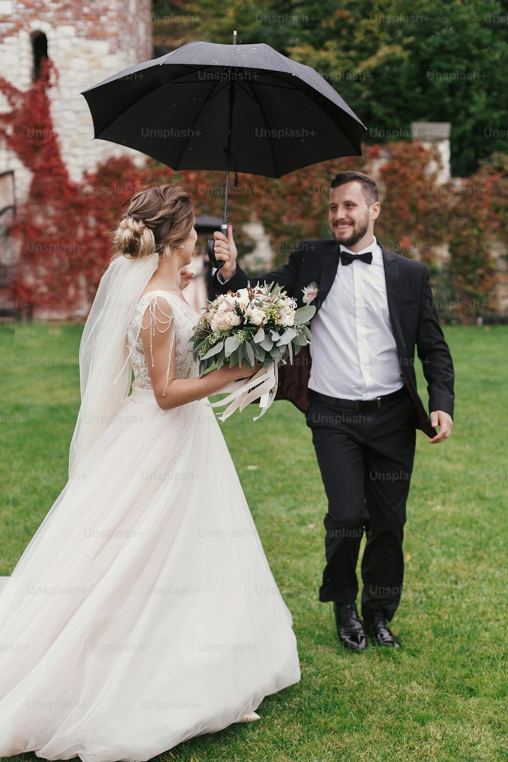 Stylish groom running under umbrella in rainy outdoor to Gorgeous bride and smiling. Sensual wedding couple having fun in rain. Romantic moments of newlyweds.